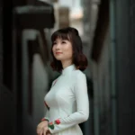 Beautiful woman wearing a white floral dress and holding a conical hat, posing in a narrow alleyway.