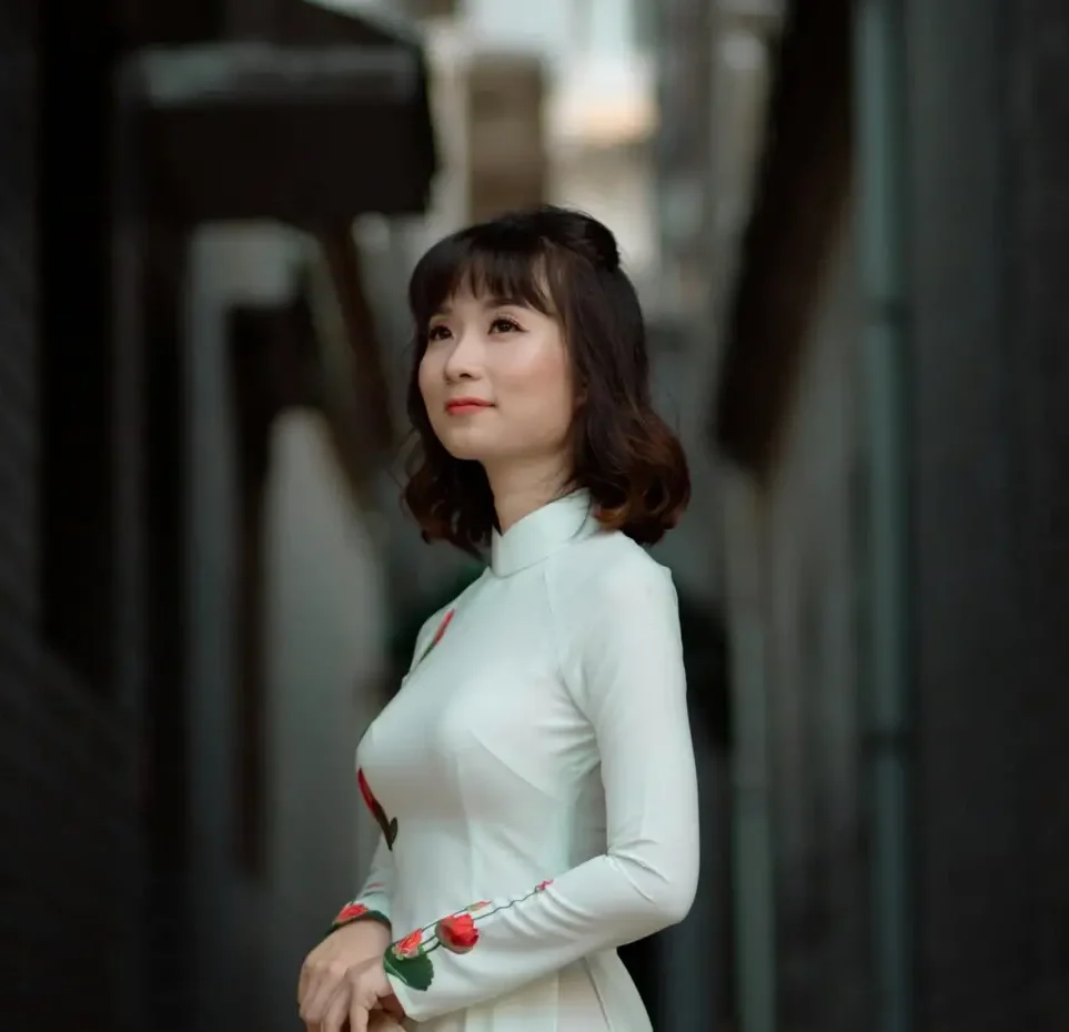 Beautiful woman wearing a white floral dress and holding a conical hat, posing in a narrow alleyway.