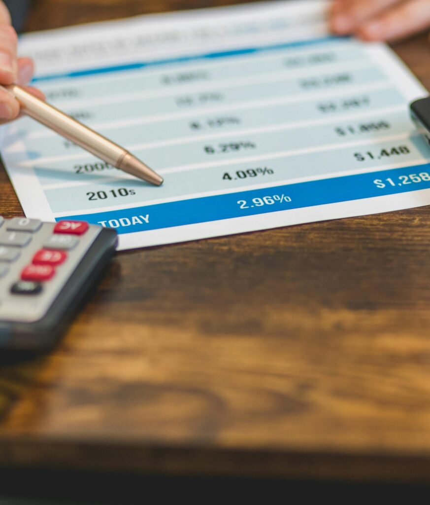 Close-up of a hand with pen analyzing financial rates on paper with a calculator and laptop nearby.